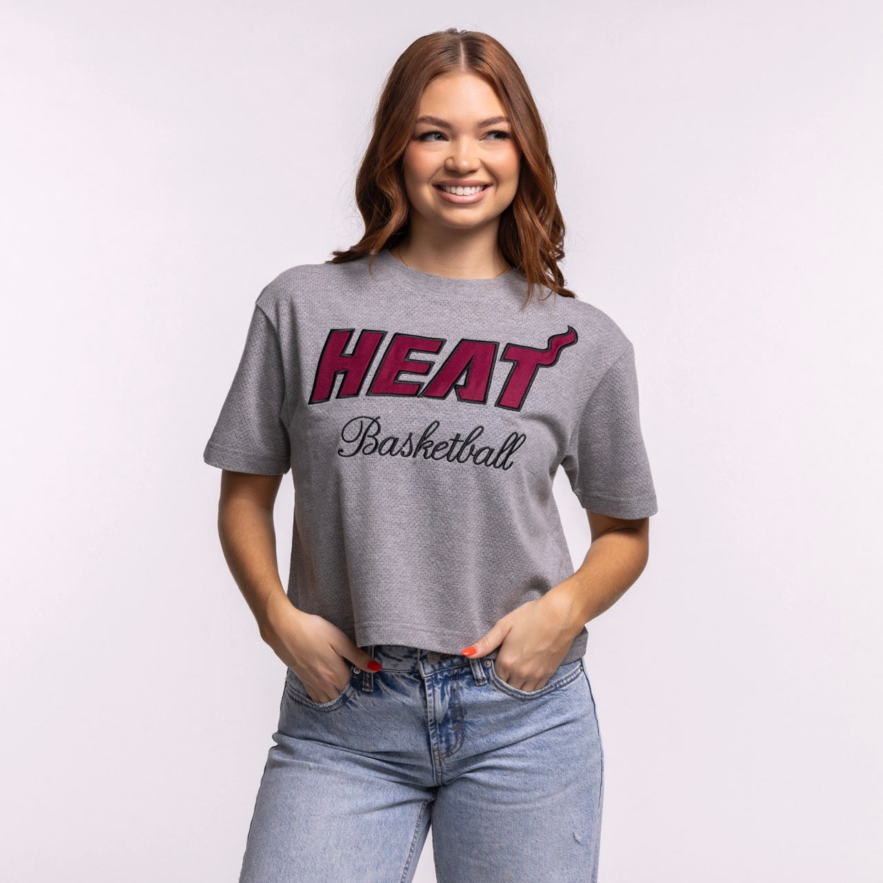 A woman smiles while wearing the '47 Brand Miami HEAT Pivot Women's Cropped Tee and light blue jeans, standing against a plain light background with her hands in her pockets to showcase the tee's relaxed cropped fit.