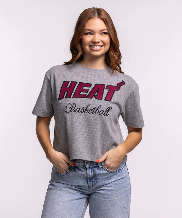 A woman smiles while wearing the '47 Brand Miami HEAT Pivot Women's Cropped Tee and light blue jeans, standing against a plain light background with her hands in her pockets to showcase the tee's relaxed cropped fit.