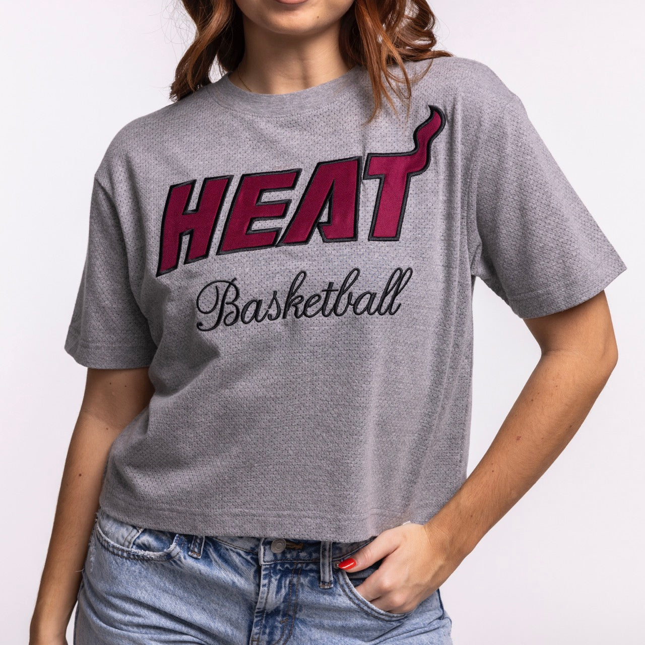 A woman wears the '47 Brand Miami HEAT Pivot Women's Cropped Tee, a relaxed gray cotton-poly blend with bold HEAT lettering and Miami Heat-inspired design, paired with high-waisted jeans. Machine wash cold.