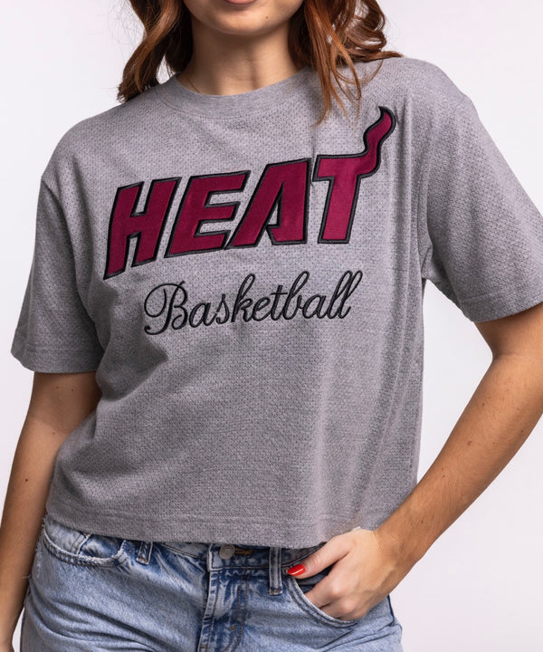 A woman smiles while wearing the '47 Brand Miami HEAT Pivot Women's Cropped Tee and light blue jeans, standing against a plain light background with her hands in her pockets to showcase the tee's relaxed cropped fit.