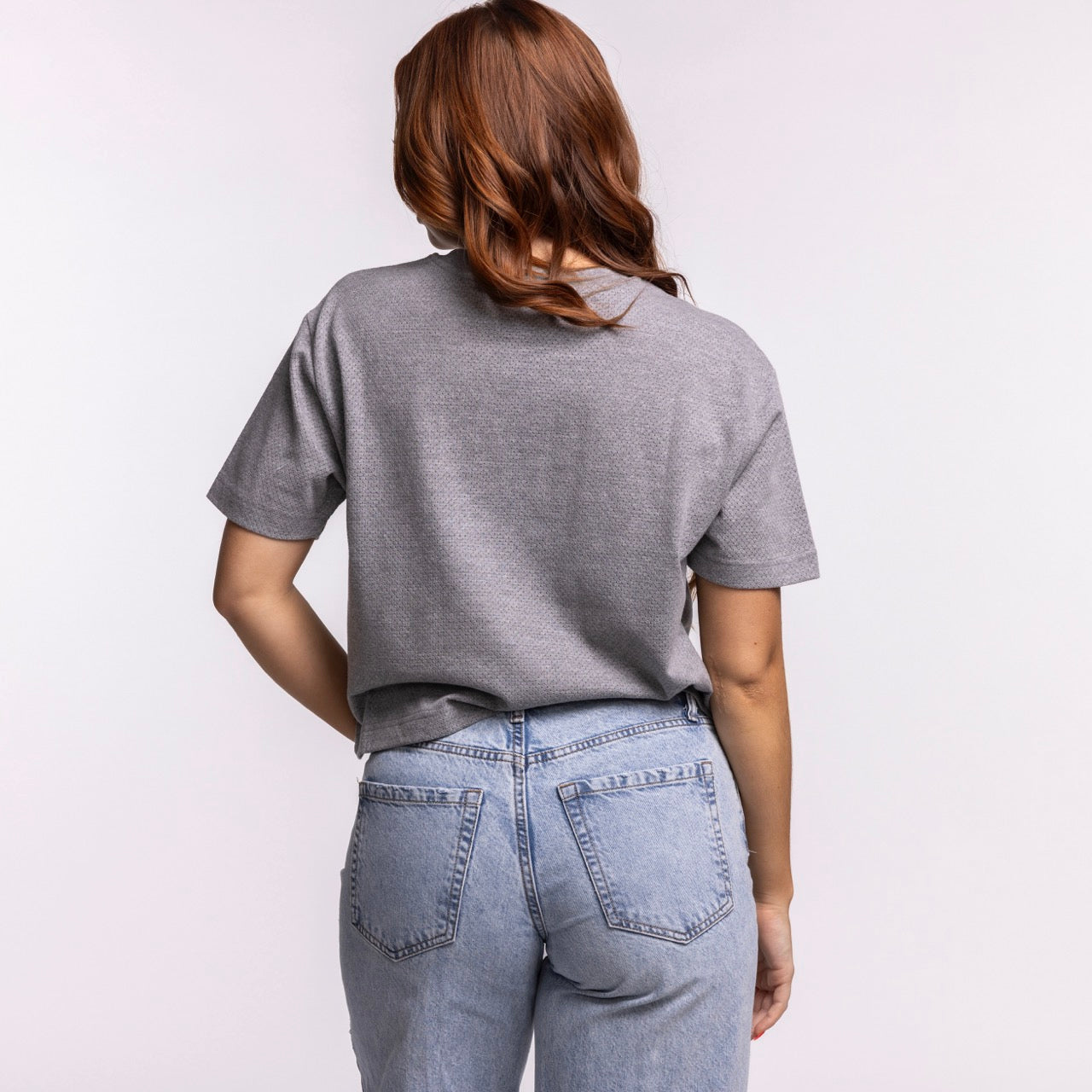 A woman with long, wavy brown hair stands with her back to the camera, wearing a '47 Brand Miami HEAT Pivot Women's Cropped Tee in gray and light blue jeans, set against a light background.