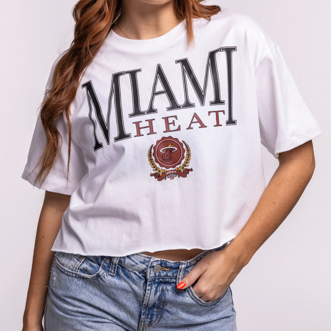 A woman wears the New Era Miami HEAT Women's Boxy Cropped Tee—a white, boxy fit tee with the team wordmark and emblem on the front—paired with light blue denim jeans.
