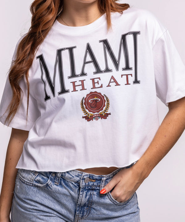 A woman with long, reddish-brown hair models the New Era Miami HEAT Women's Boxy Cropped Tee in white, showcasing the bold HEAT wordmark, paired with blue jeans, standing against a plain light background and smiling slightly.