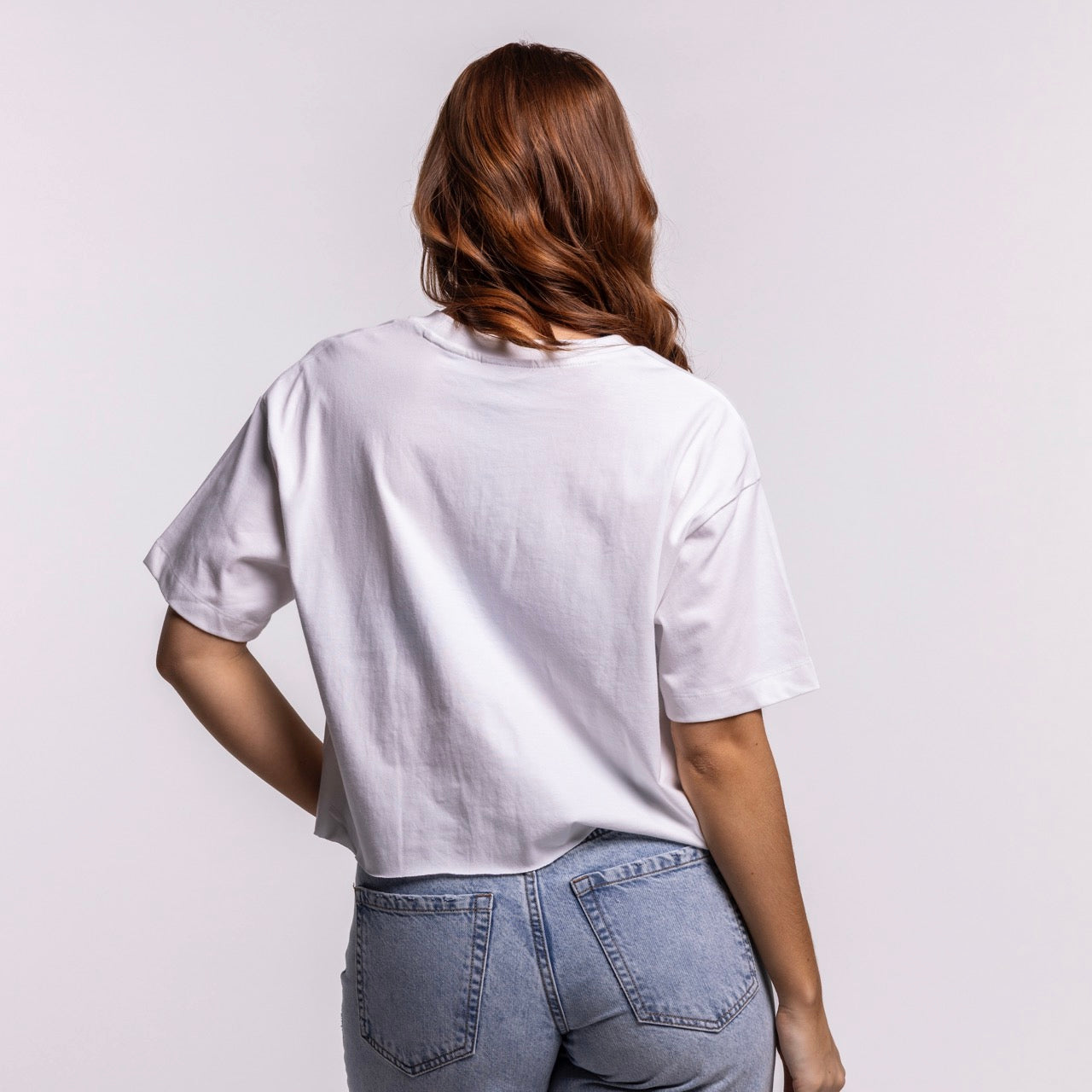 A woman with long, wavy, auburn hair faces away from the camera, wearing the New Era Miami HEAT Women's Boxy Cropped Tee and light blue jeans against a plain light background.