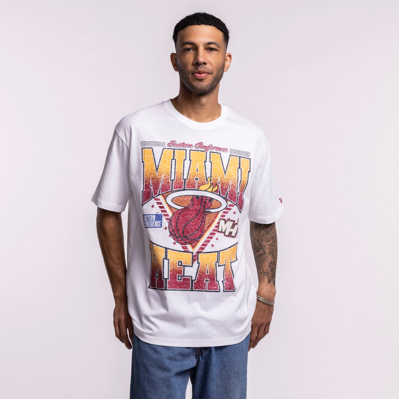 A man, smiling with tattoos on his left forearm, wears a New Era Miami HEAT Waterbased Tee in optic white and blue jeans, proudly displaying Miami HEAT spirit against a plain background.