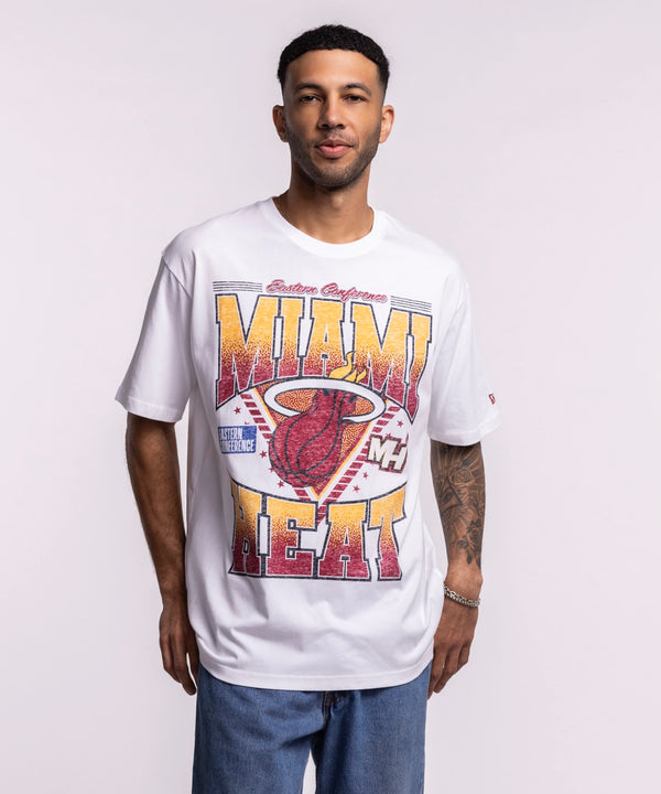 A man, smiling with tattoos on his left forearm, wears a New Era Miami HEAT Waterbased Tee in optic white and blue jeans, proudly displaying Miami HEAT spirit against a plain background.