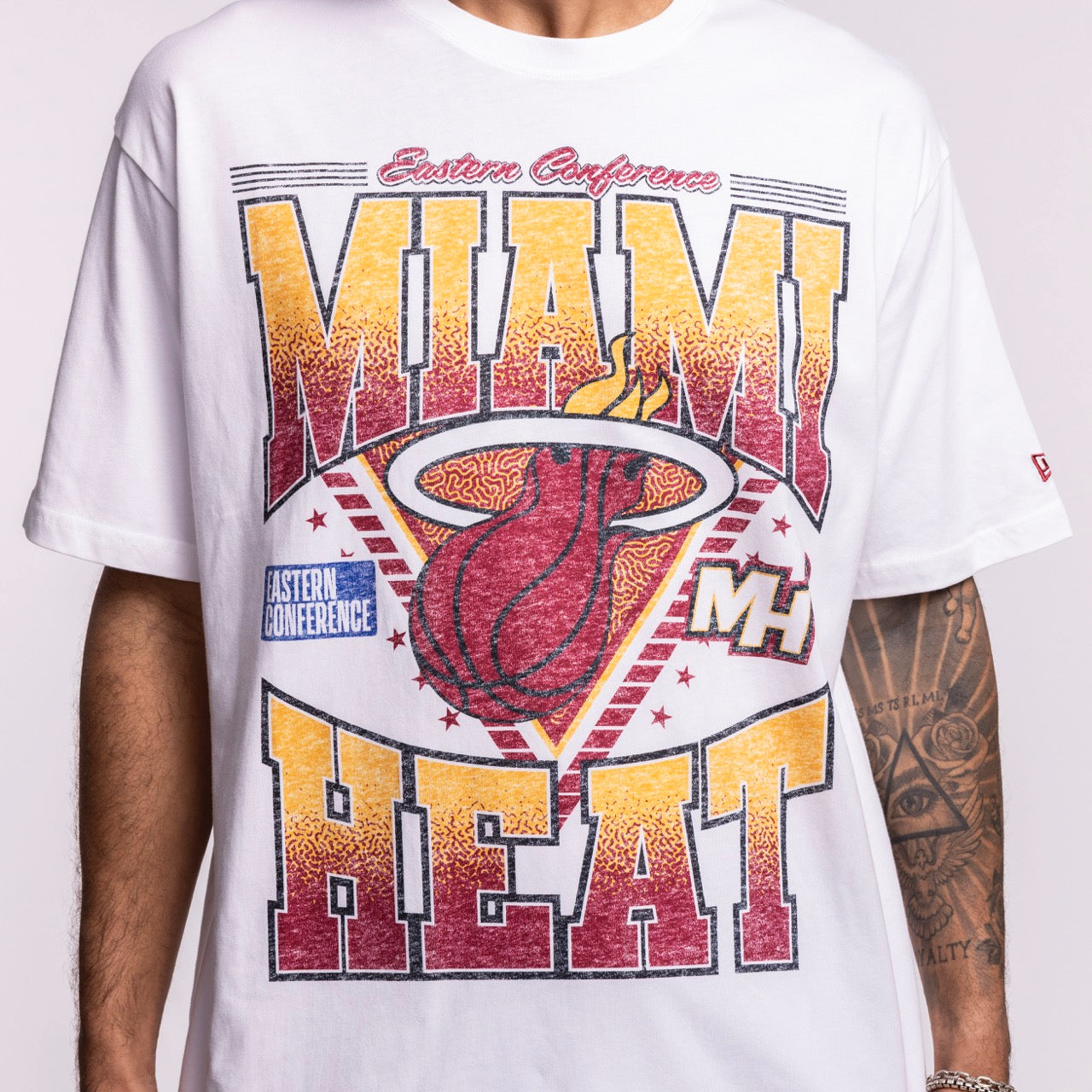 A person wears the New Era Miami HEAT Waterbased Tee, an optic white shirt with a bold HEAT graphic, Miami HEAT logo, 