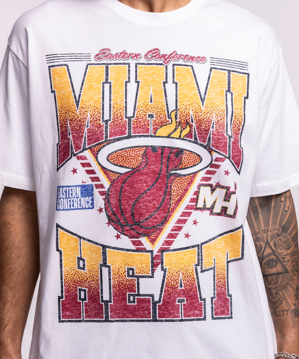 A man, smiling with tattoos on his left forearm, wears a New Era Miami HEAT Waterbased Tee in optic white and blue jeans, proudly displaying Miami HEAT spirit against a plain background.