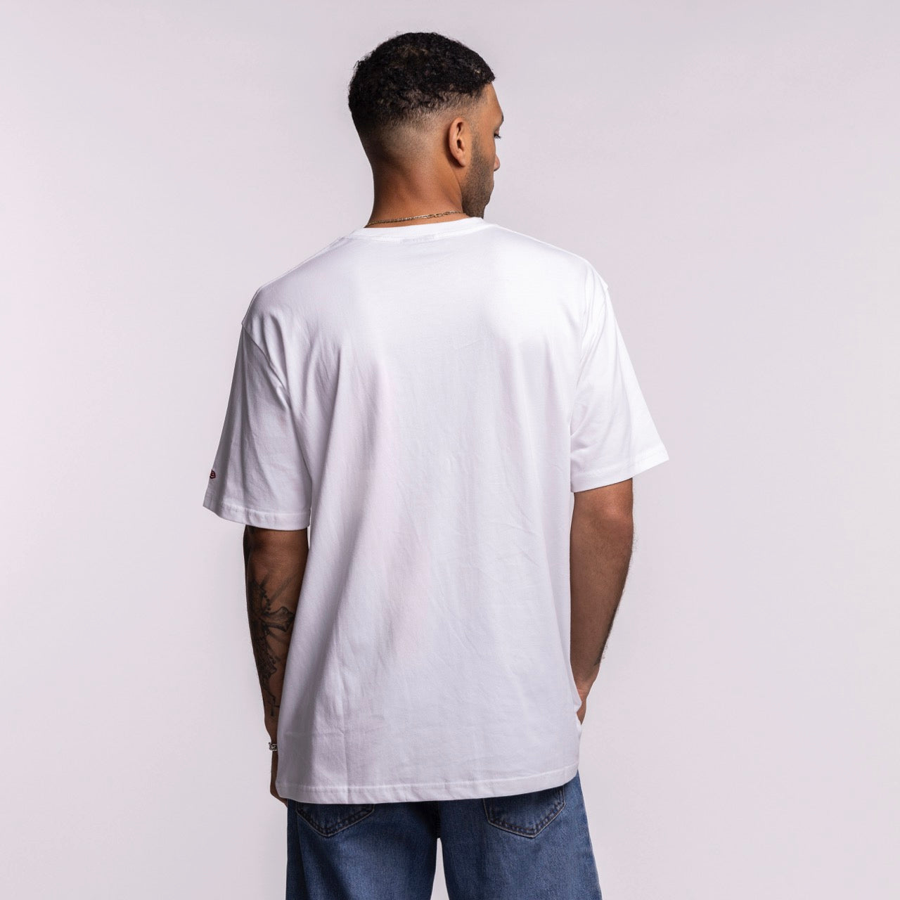 A man stands facing away from the camera, wearing the New Era Miami HEAT Waterbased Tee and blue jeans. His arms are relaxed by his sides, showing tattoos on his left forearm, against a light gray background.