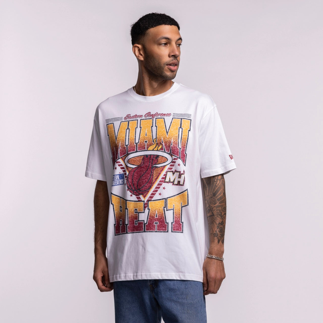 A man in a New Era Miami HEAT Waterbased Tee with a bold, screen-printed HEAT graphic and blue jeans stands against a light background, looking to his left.