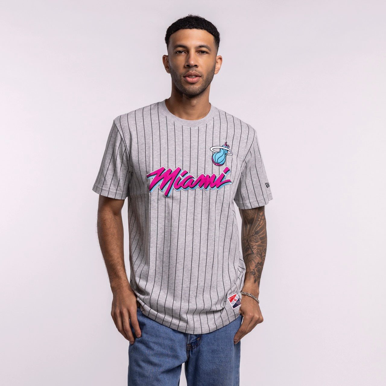 A man stands against a plain background wearing the New Era Miami HEAT Original Vice Nights Striped Tee—gray pinstripe with bright Miami/basketball logo—and blue jeans. He has a visible arm tattoo. Standard fit; machine wash recommended.