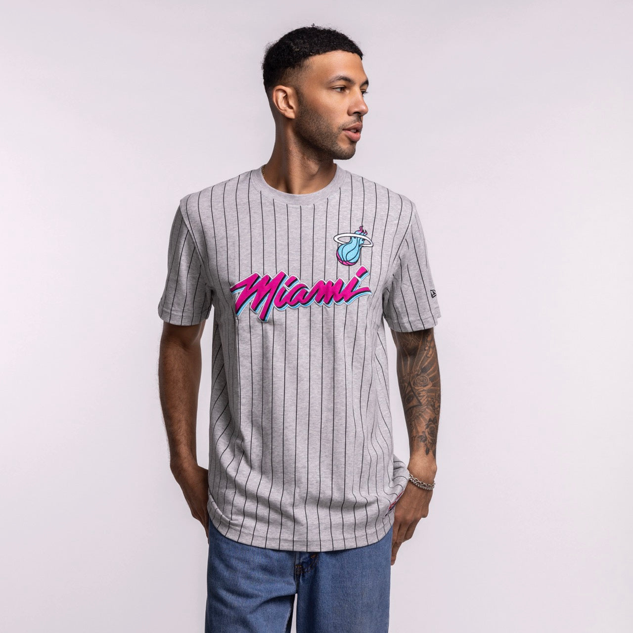 A man wears the New Era Miami HEAT Original Vice Nights Striped Tee, a gray pinstriped cotton-polyester shirt with pink and blue lettering. He stands against a plain background, looking left, hands at his sides. Standard fit by New Era.
