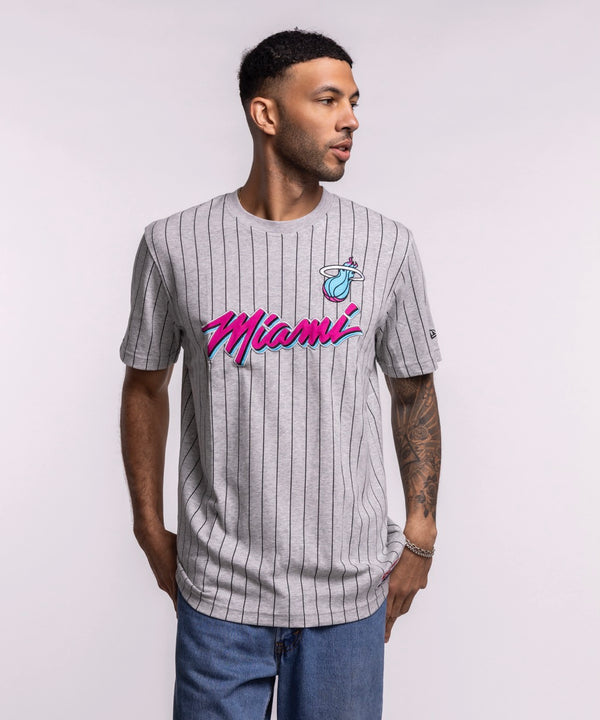 A man wears the New Era Miami HEAT Original Vice Nights Striped Tee, a gray pinstriped cotton-polyester shirt with pink and blue lettering. He stands against a plain background, looking left, hands at his sides. Standard fit by New Era.