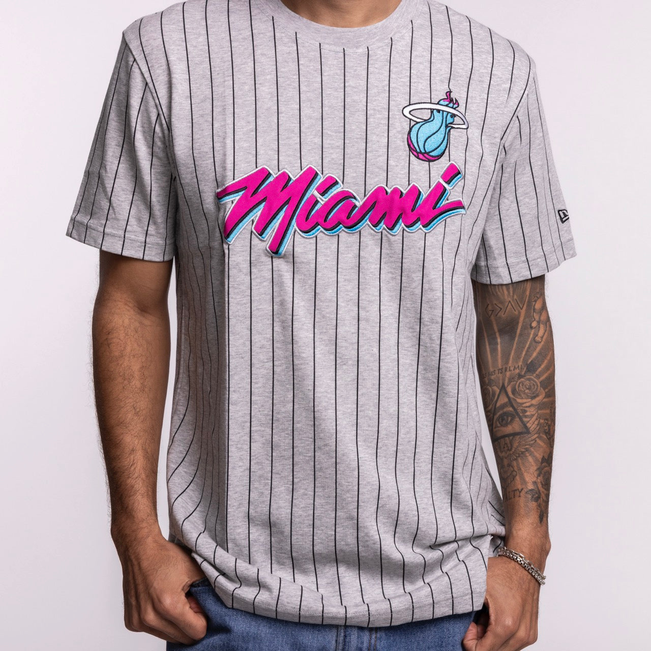 A person wears the New Era Miami HEAT Original Vice Nights Striped Tee by New Era—a gray pinstriped t-shirt with “Miami” in pink and blue script and a flaming basketball logo—made from soft cotton-polyester, against a plain light background.