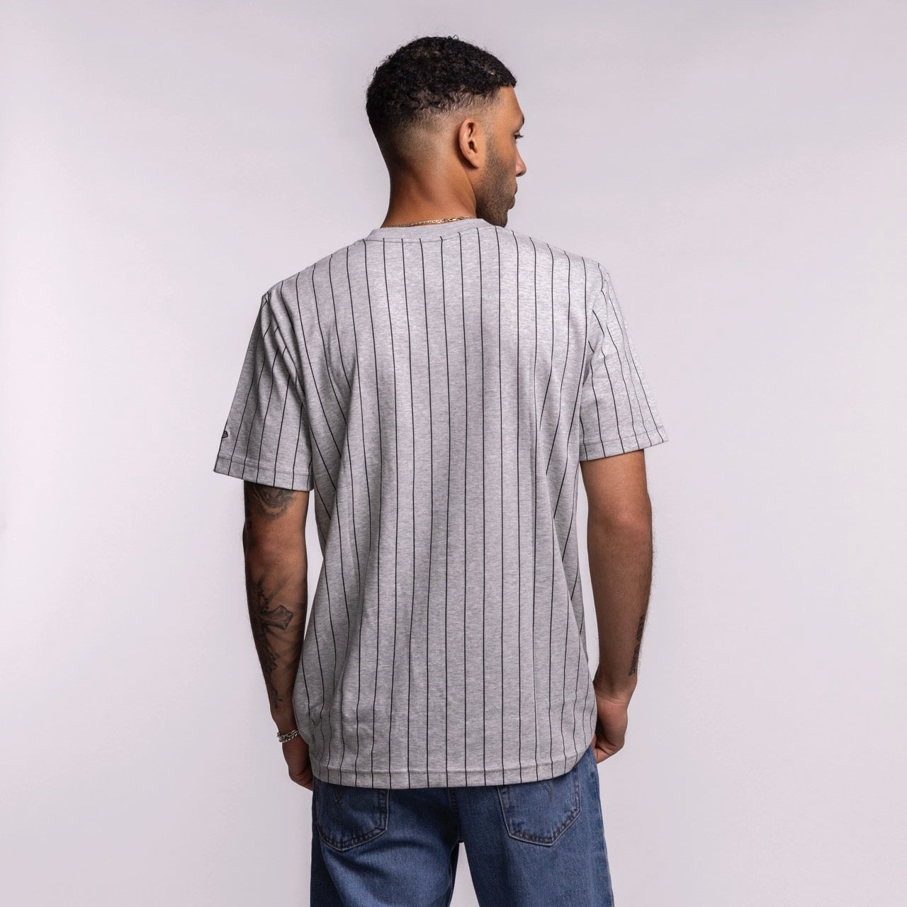 A man with short curly hair, seen from behind, wears a New Era Miami HEAT Original Vice Nights Striped Tee—standard fit, light gray with thin black vertical stripes—and blue jeans. The cotton-poly blend shirt stands out against a plain, light background.