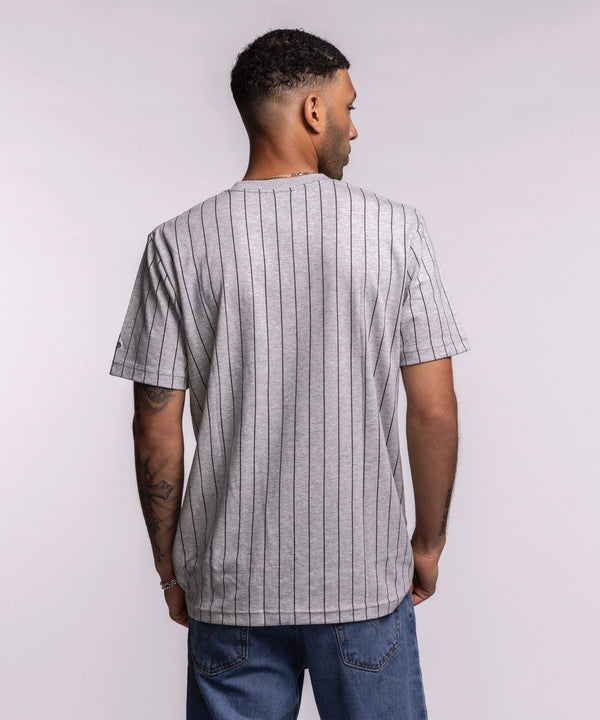 A man wears the New Era Miami HEAT Original Vice Nights Striped Tee, a gray pinstriped cotton-polyester shirt with pink and blue lettering. He stands against a plain background, looking left, hands at his sides. Standard fit by New Era.