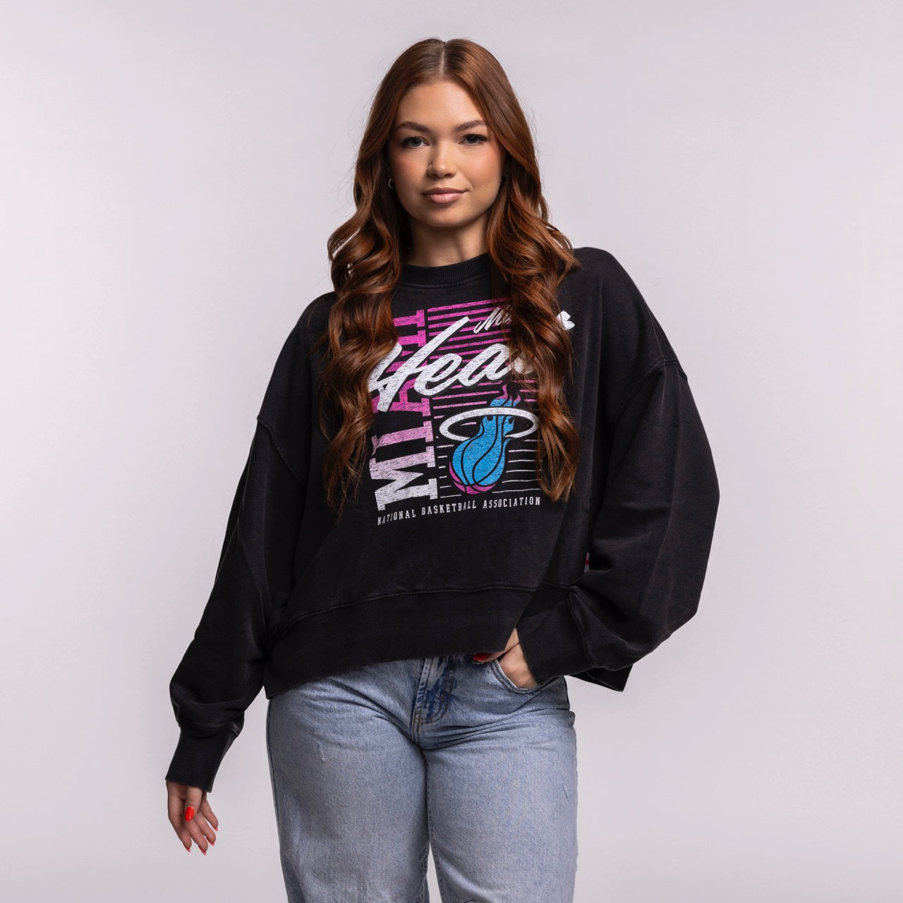 A woman with long, wavy brown hair wears the New Era Miami HEAT Original Vice Nights Washed Women's Crewneck with a relaxed fit and light blue jeans, standing against a plain background and smiling slightly with one hand in her pocket.