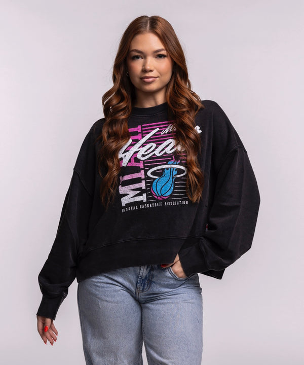A woman with long, wavy brown hair wears the New Era Miami HEAT Original Vice Nights Washed Women's Crewneck with a relaxed fit and light blue jeans, standing against a plain background and smiling slightly with one hand in her pocket.