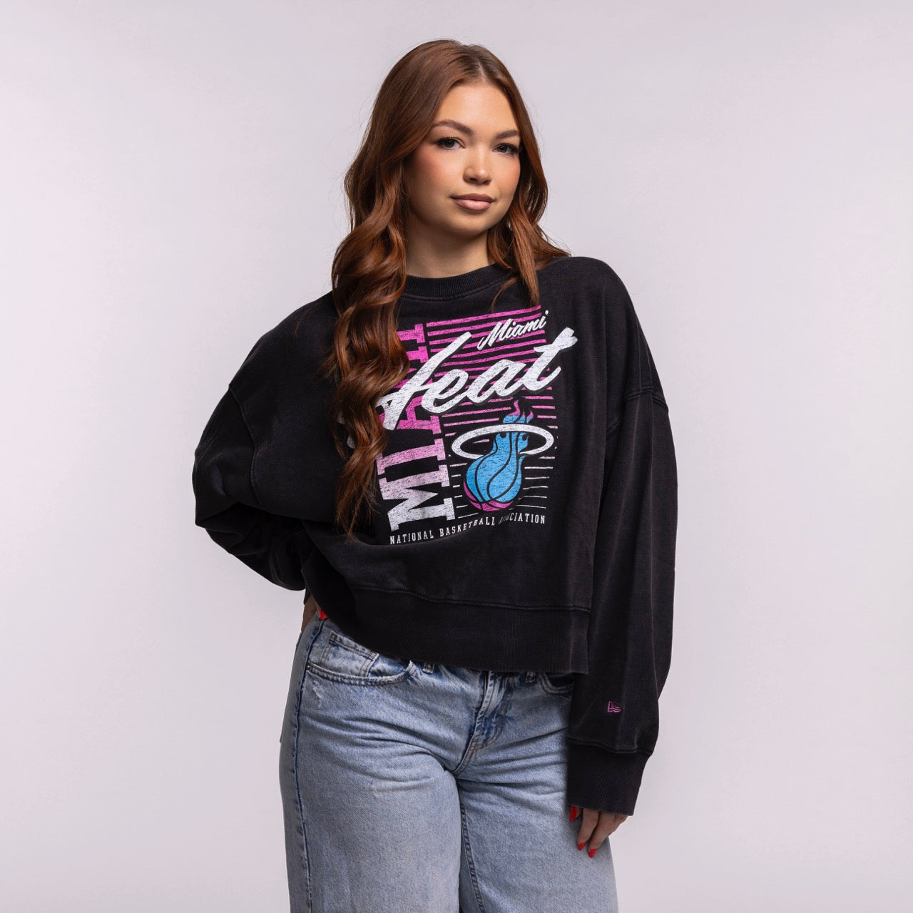 A young woman with long brown hair wears the New Era Miami HEAT Original Vice Nights Washed Women's Crewneck and light blue jeans. She stands against a plain light gray background, hand on her hip and looking relaxed.