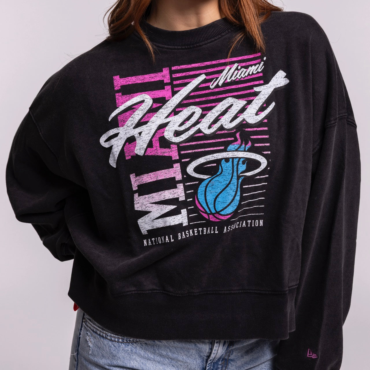 A person in a New Era Miami HEAT Original Vice Nights Washed Women's Crewneck—black with a bold retro design in white, pink, and teal—styled with light blue jeans. The person’s face isn’t visible.
