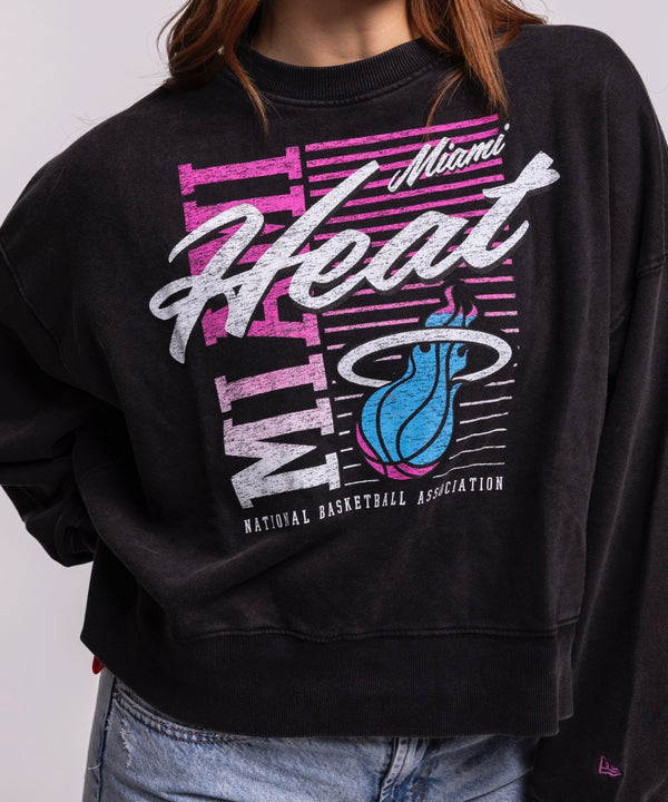 A woman with long, wavy brown hair wears the New Era Miami HEAT Original Vice Nights Washed Women's Crewneck with a relaxed fit and light blue jeans, standing against a plain background and smiling slightly with one hand in her pocket.