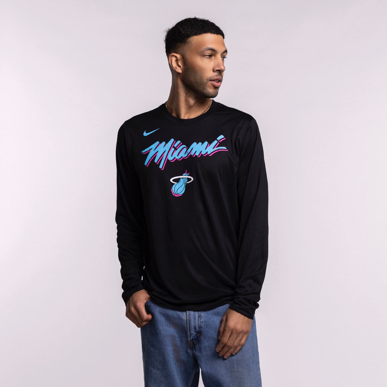 A man wearing a men's size L Nike Miami HEAT Original Vice Nights Legend Long Sleeve Tee in black with pink and blue lettering and blue jeans stands against a plain light background, looking to his left with one hand in his pocket.