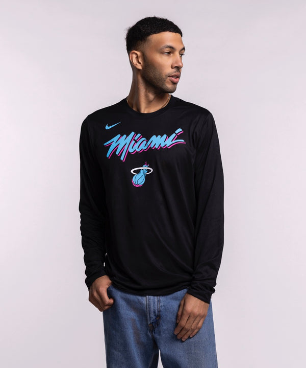 A man wearing a men's size L Nike Miami HEAT Original Vice Nights Legend Long Sleeve Tee in black with pink and blue lettering and blue jeans stands against a plain light background, looking to his left with one hand in his pocket.