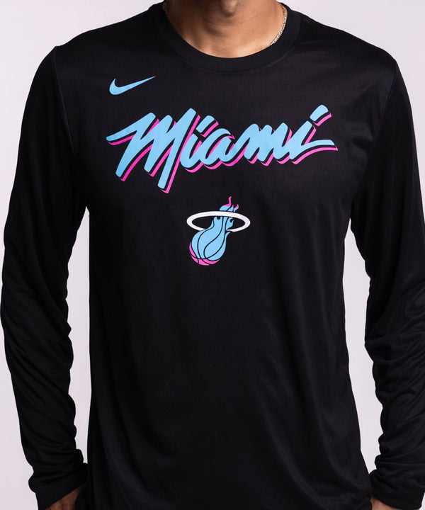 A man wearing a men's size L Nike Miami HEAT Original Vice Nights Legend Long Sleeve Tee in black with pink and blue lettering and blue jeans stands against a plain light background, looking to his left with one hand in his pocket.