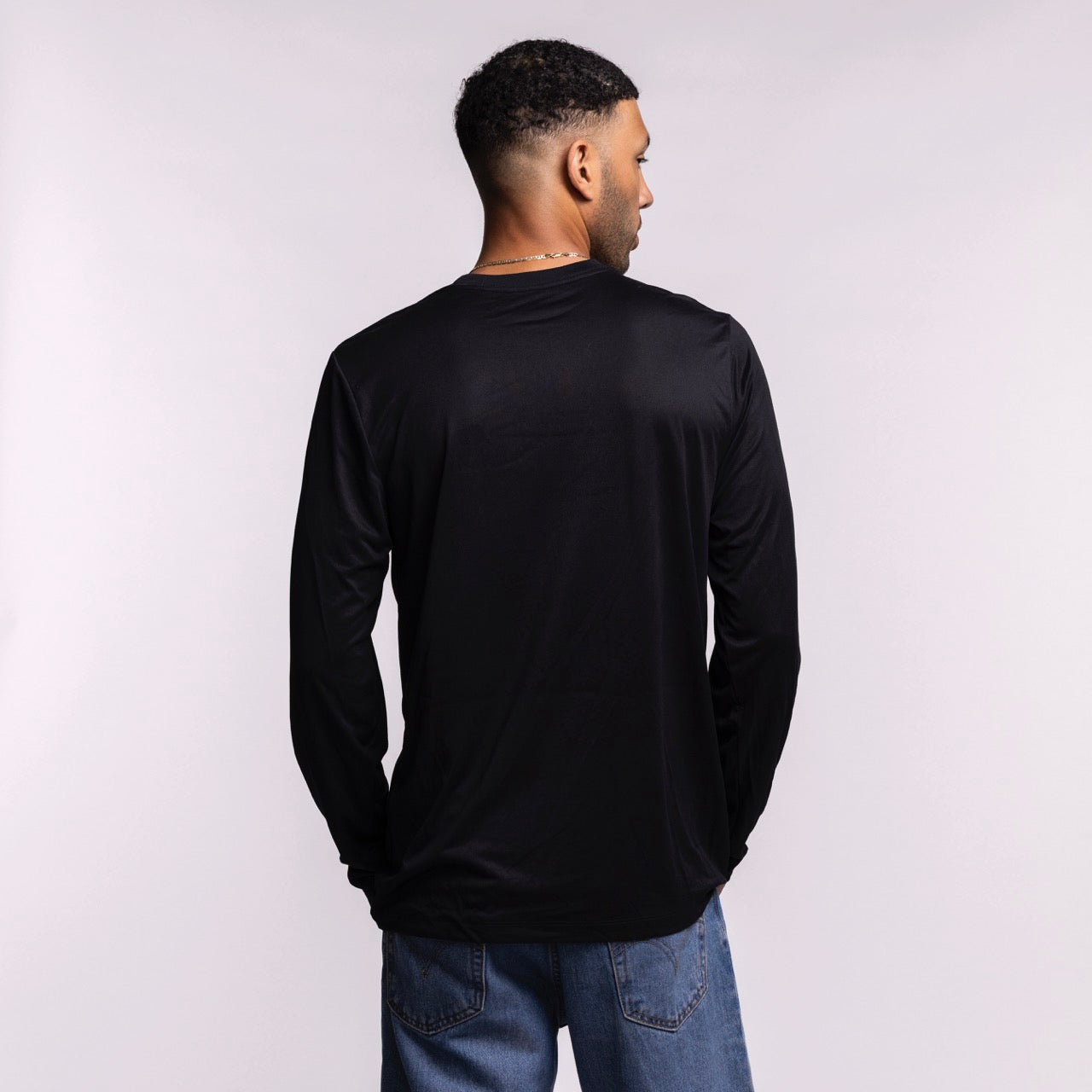 A man stands facing away from the camera, wearing a men's size L Nike Miami HEAT Original Vice Nights Legend Long Sleeve Tee in black and blue jeans, set against a plain light background.