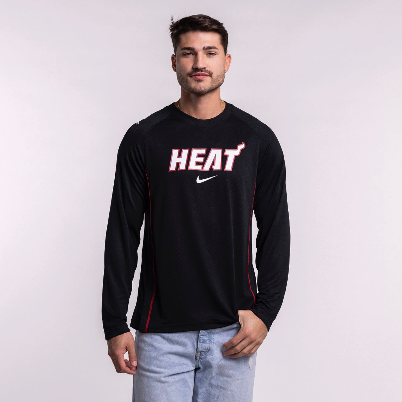 A man in a Nike Miami HEAT 2025-26 On-Court Long Sleeve Shooting Shirt and light blue jeans stands against a plain light background, looking at the camera with a neutral expression.