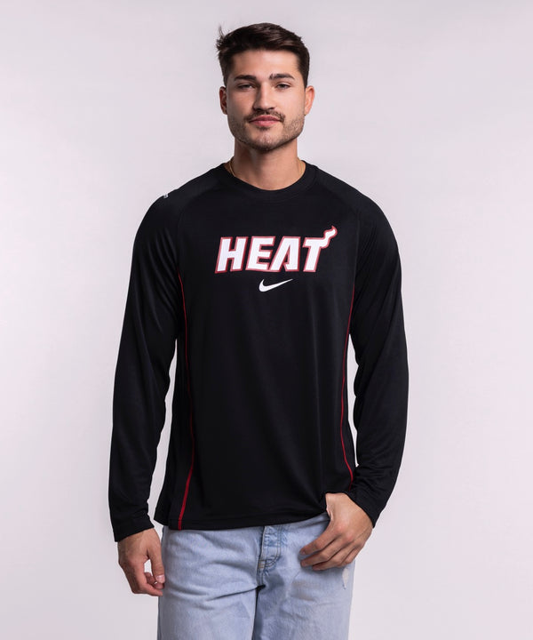 A man in a Nike Miami HEAT 2025-26 On-Court Long Sleeve Shooting Shirt and light blue jeans stands against a plain light background, looking at the camera with a neutral expression.