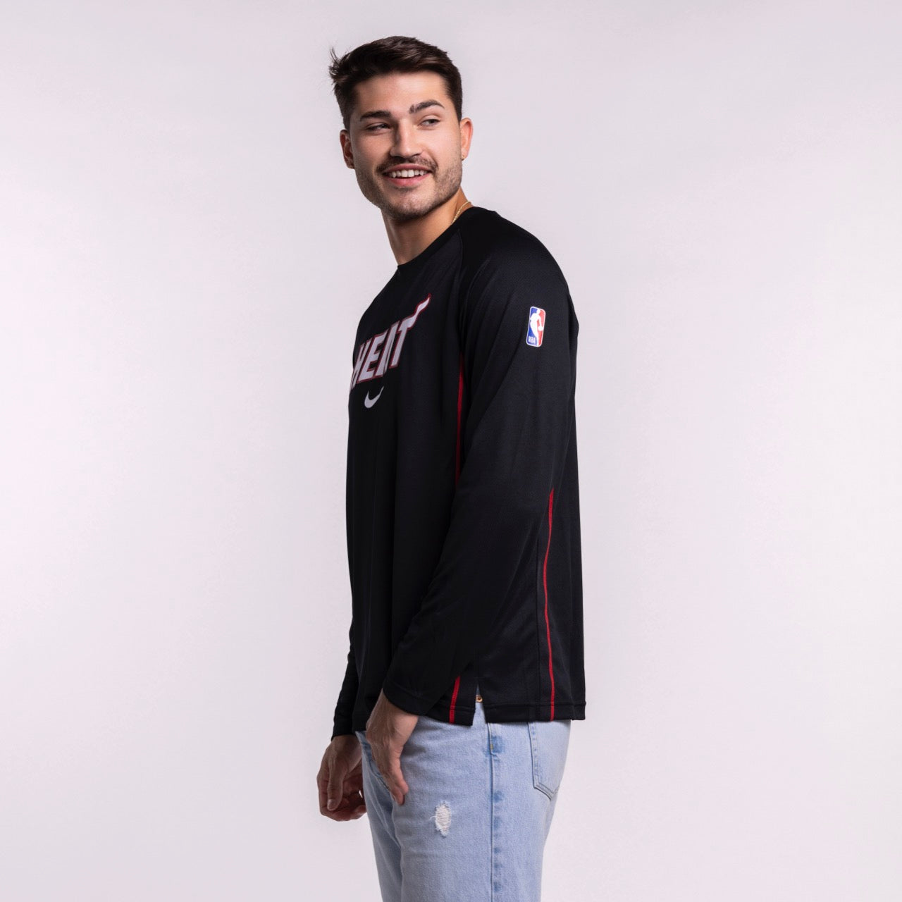 A young man with short dark hair and a mustache is wearing the Nike Miami HEAT 2025-26 On-Court Long Sleeve Shooting Shirt, standing sideways and smiling in light blue jeans against a plain light background.