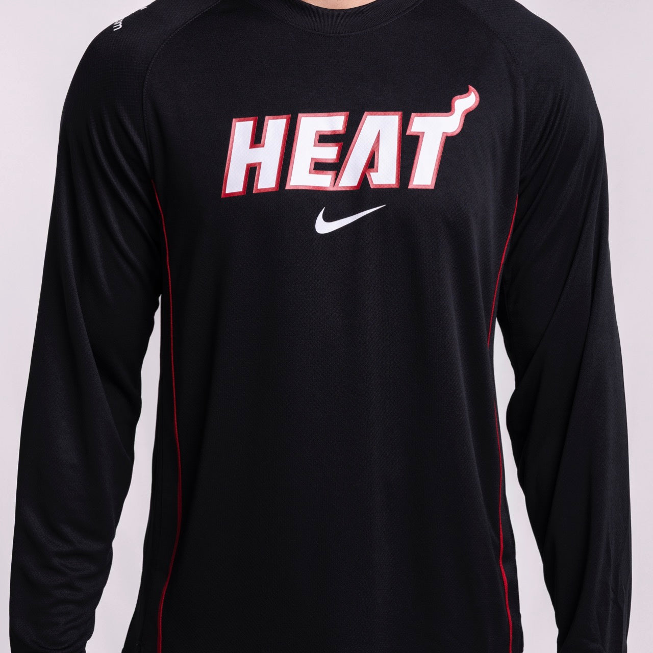 A person wears the Nike Miami HEAT 2025-26 On-Court Long Sleeve Shooting Shirt, featuring bold white and red team lettering on the front. This moisture-wicking Dri-FIT shirt from Nike is shown from the torso up.