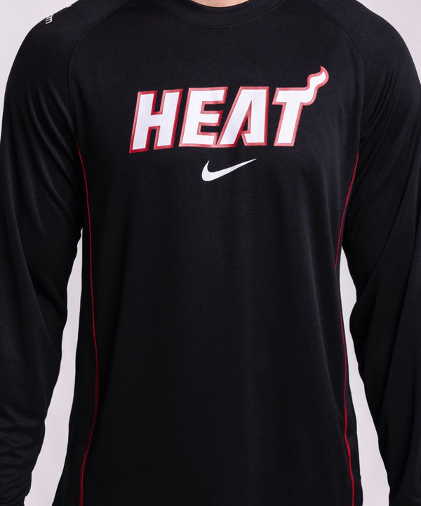 A man in a Nike Miami HEAT 2025-26 On-Court Long Sleeve Shooting Shirt and light blue jeans stands against a plain light background, looking at the camera with a neutral expression.