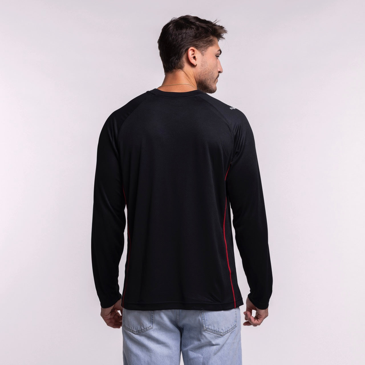 A man with short brown hair stands with his back to the camera, wearing a Nike Miami HEAT 2025-26 On-Court Long Sleeve Shooting Shirt and light blue jeans against a plain light background.