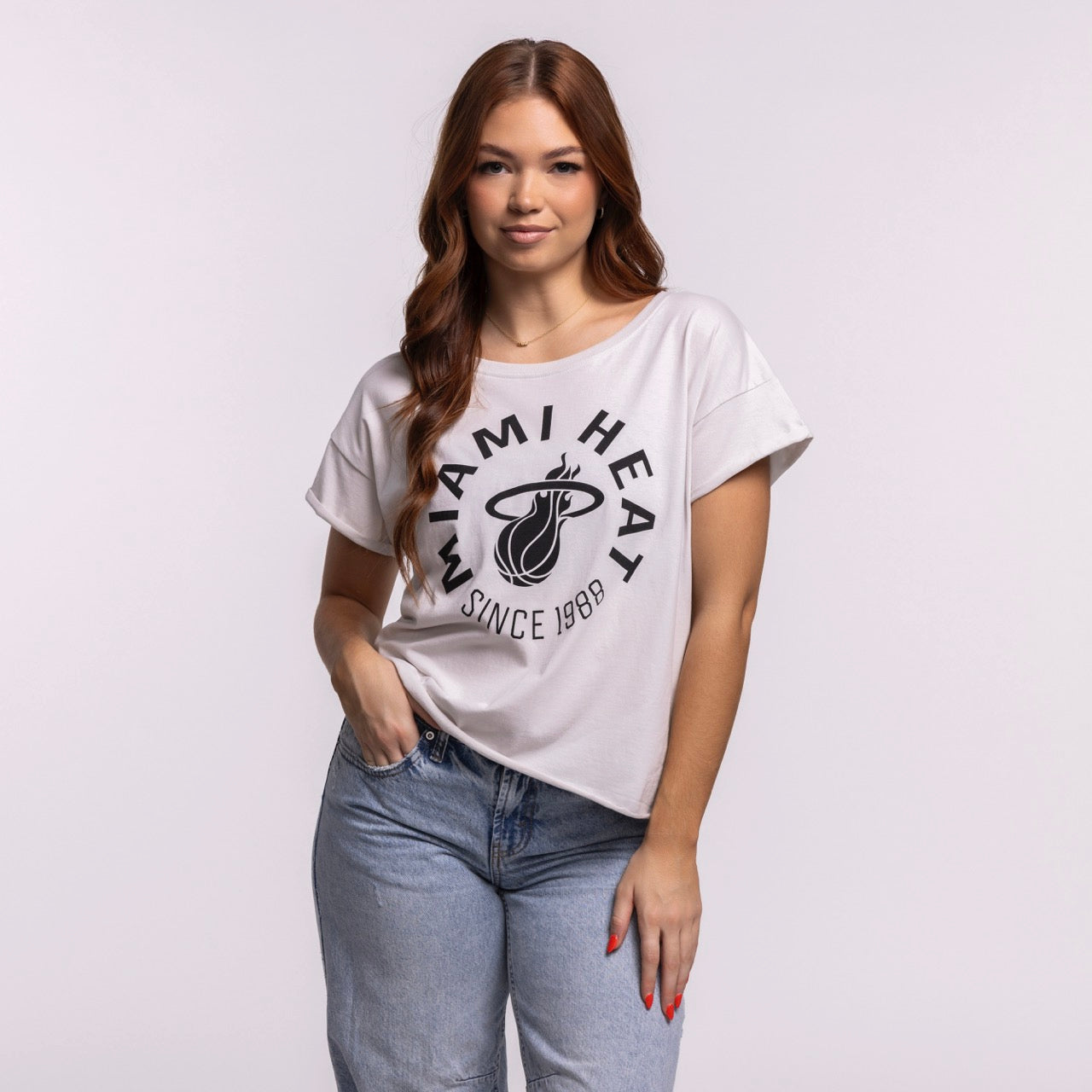 A woman with long brown hair stands against a plain background, wearing the Sportiqe Miami HEAT 1988 Basketball Women's Tee in a relaxed cotton fit and light blue jeans, one hand in her pocket and a slight smile on her face.