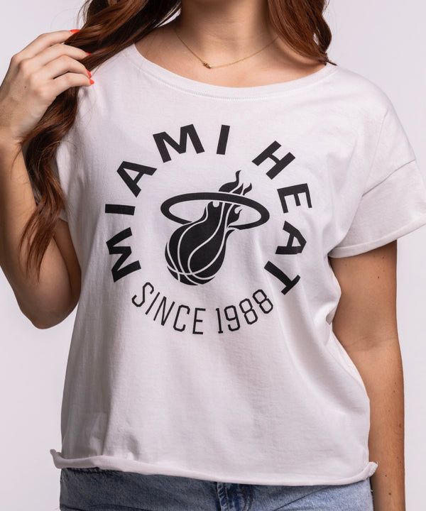 A woman with long brown hair stands against a plain background, wearing the Sportiqe Miami HEAT 1988 Basketball Women's Tee in a relaxed cotton fit and light blue jeans, one hand in her pocket and a slight smile on her face.