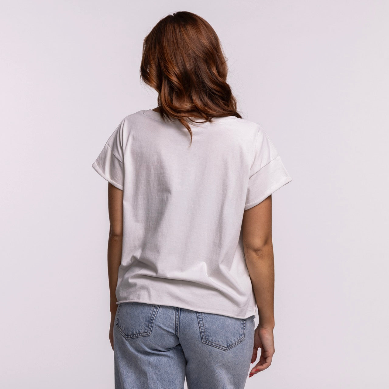 A woman with wavy, shoulder-length brown hair stands with her back to the camera, wearing the Sportiqe Miami HEAT 1988 Basketball Women's Tee and light blue jeans against a plain light background.