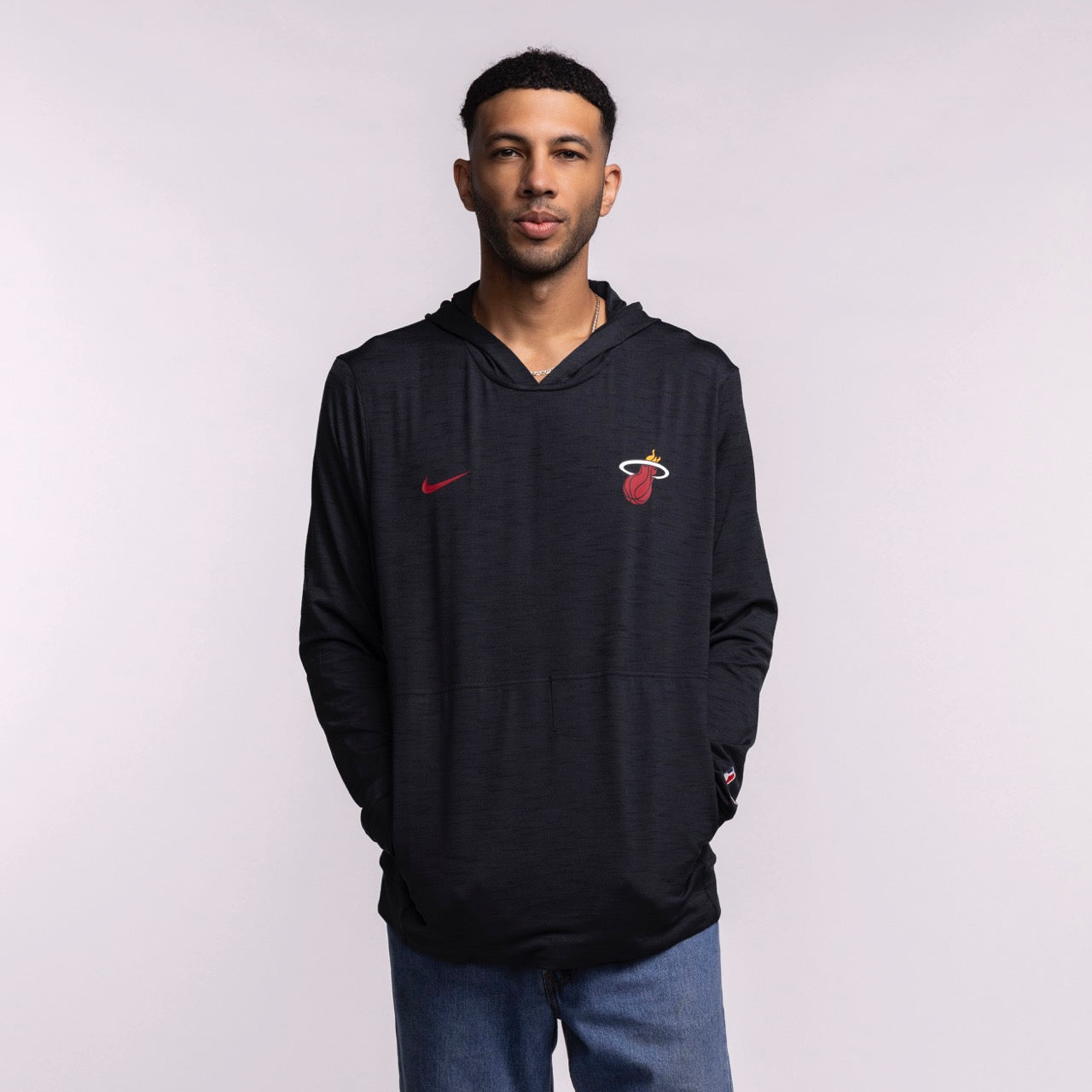A man faces forward in a Nike Miami HEAT Restore Pullover Hoodie featuring sweat-wicking tech and a red Nike logo, paired with blue jeans against a plain light background.