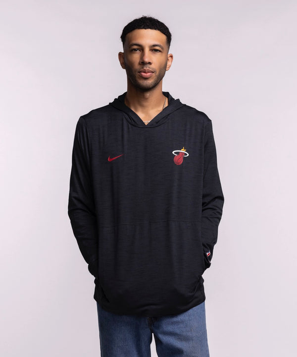 A man faces forward in a Nike Miami HEAT Restore Pullover Hoodie featuring sweat-wicking tech and a red Nike logo, paired with blue jeans against a plain light background.