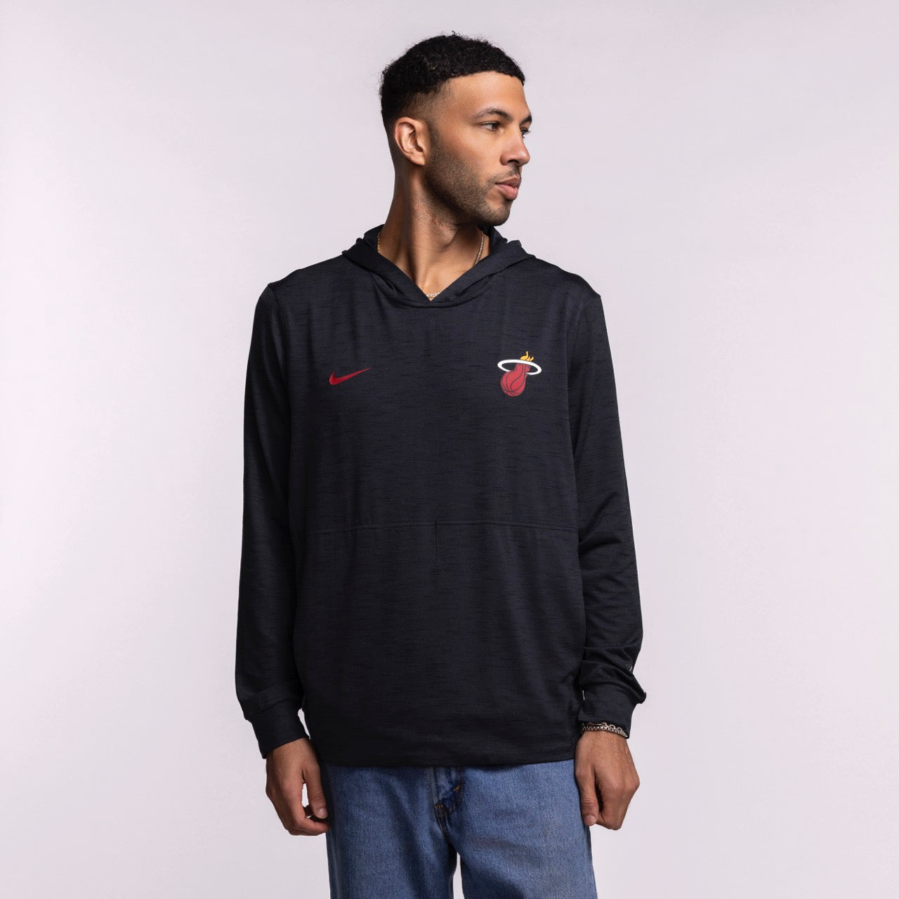 A man in a black Nike Miami HEAT Restore Pullover Hoodie with sweat-wicking tech and a red Nike logo stands against a plain light background, looking left. He pairs the hoodie with blue jeans.