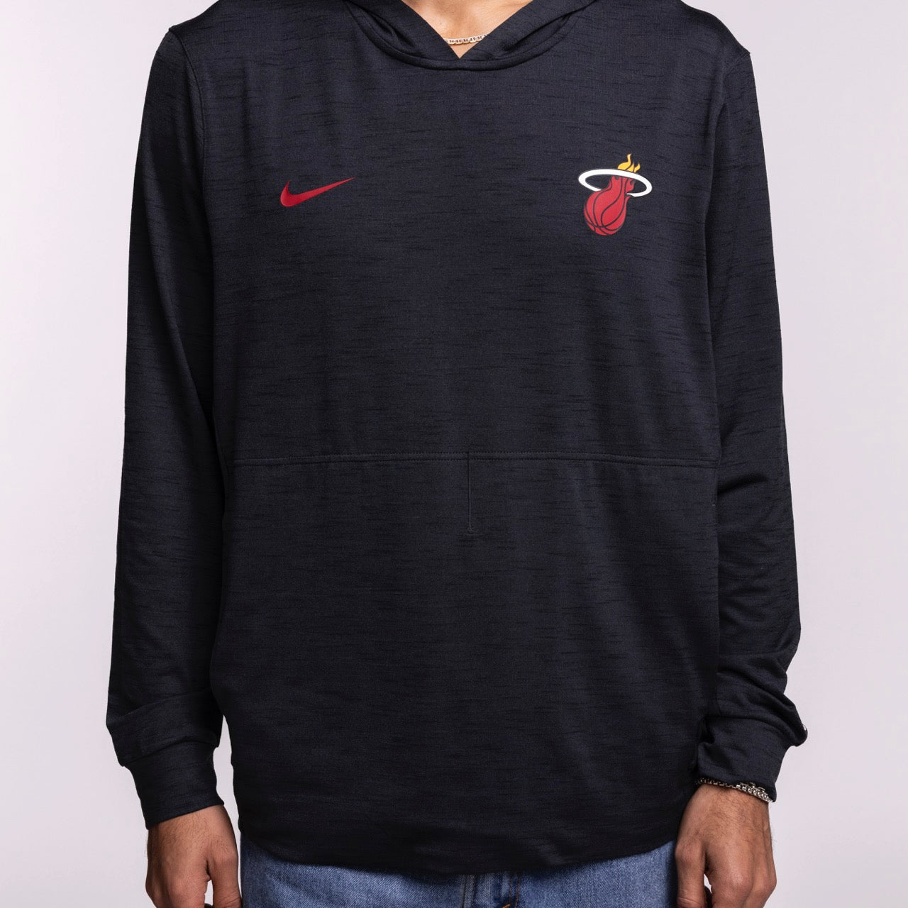 A person wears the Nike Miami HEAT Restore Pullover Hoodie, featuring a black design, red Nike logo on the left, Miami HEAT flaming basketball logo on the right, long sleeves, front pocket, and sweat-wicking comfort.
