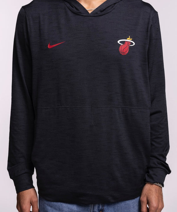 A man faces forward in a Nike Miami HEAT Restore Pullover Hoodie featuring sweat-wicking tech and a red Nike logo, paired with blue jeans against a plain light background.