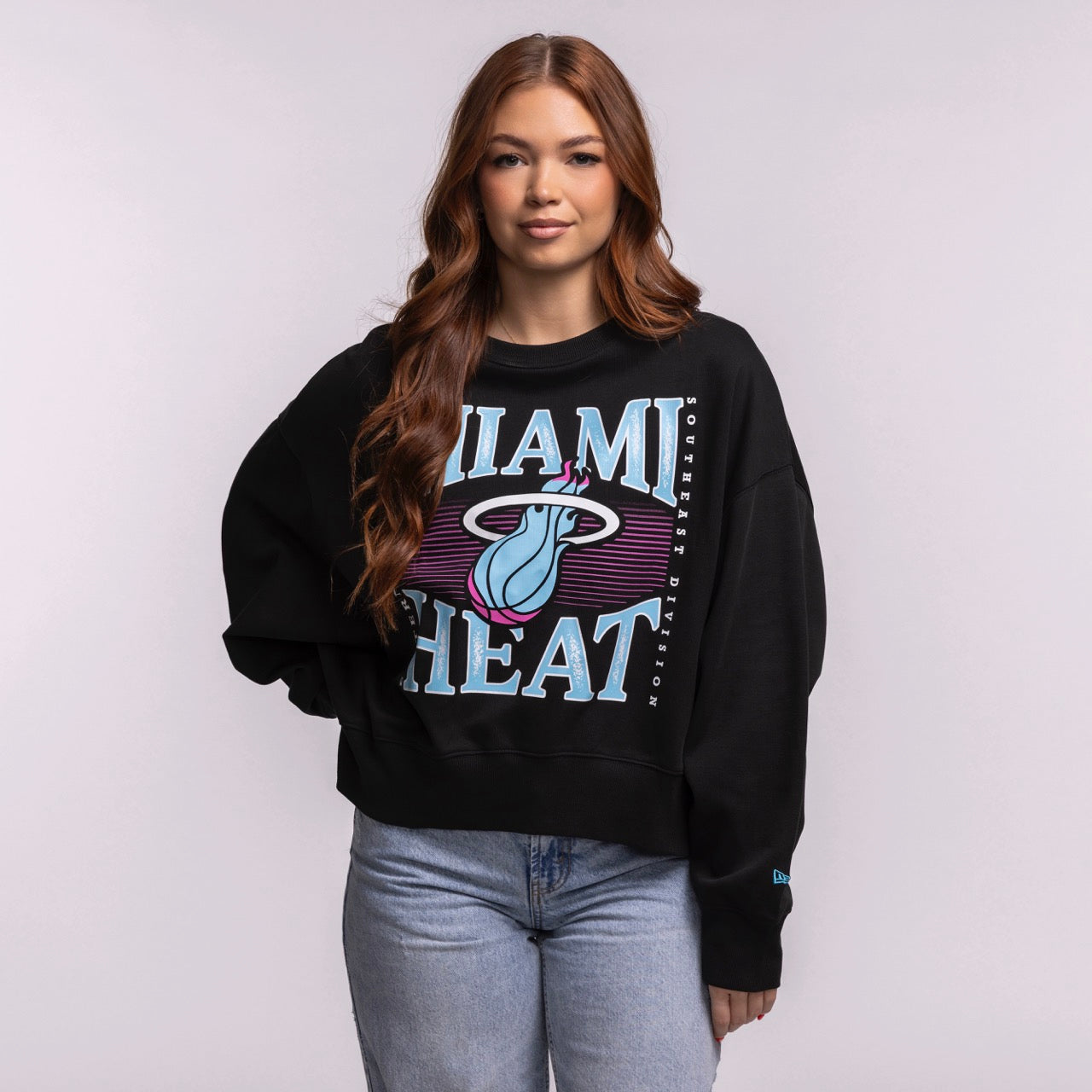 A woman with long brown hair wears the New Era Miami HEAT Original Vice Nights Wordmark Women's Crewneck in black with an oversized cropped fit and light blue jeans, standing against a plain background facing the camera.