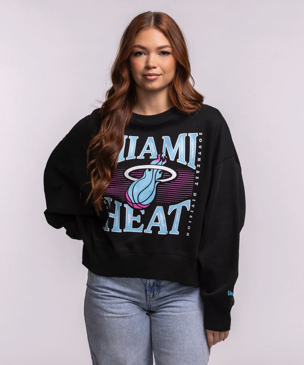 A woman with long brown hair wears the New Era Miami HEAT Original Vice Nights Wordmark Women's Crewneck in black with an oversized cropped fit and light blue jeans, standing against a plain background facing the camera.