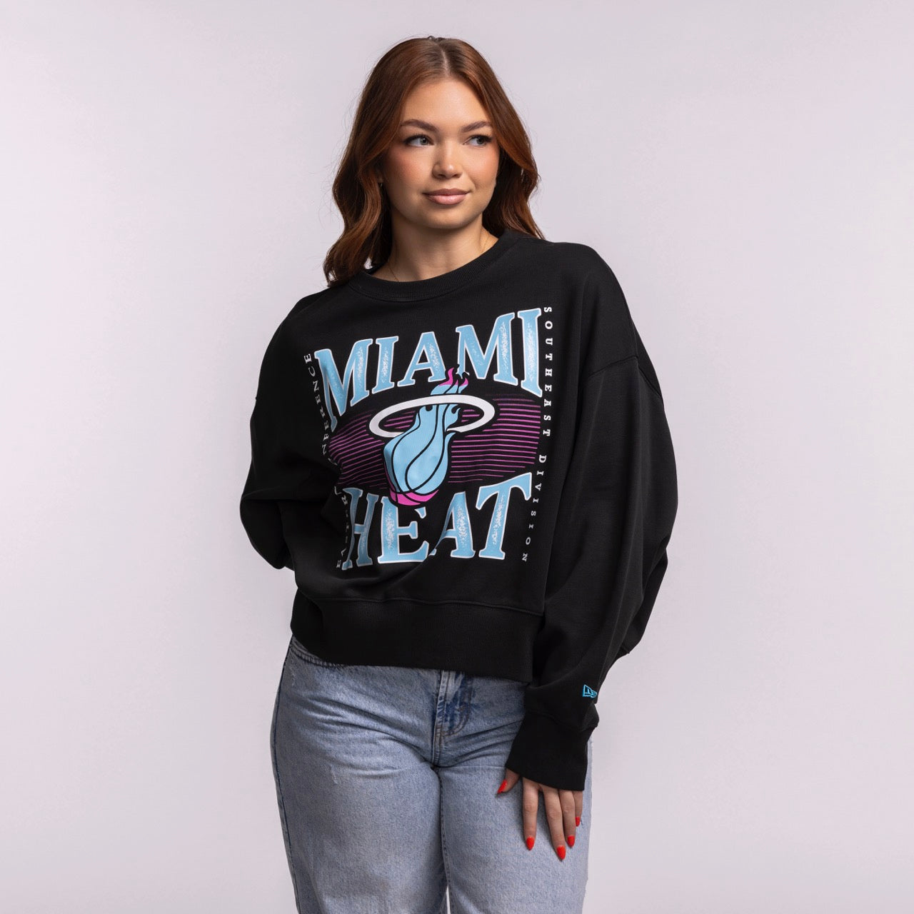 A woman with long brown hair wears a New Era Miami HEAT Original Vice Nights Wordmark Women's Crewneck—an oversized black sweatshirt with blue and pink lettering—paired with light blue jeans, standing against a plain light background.