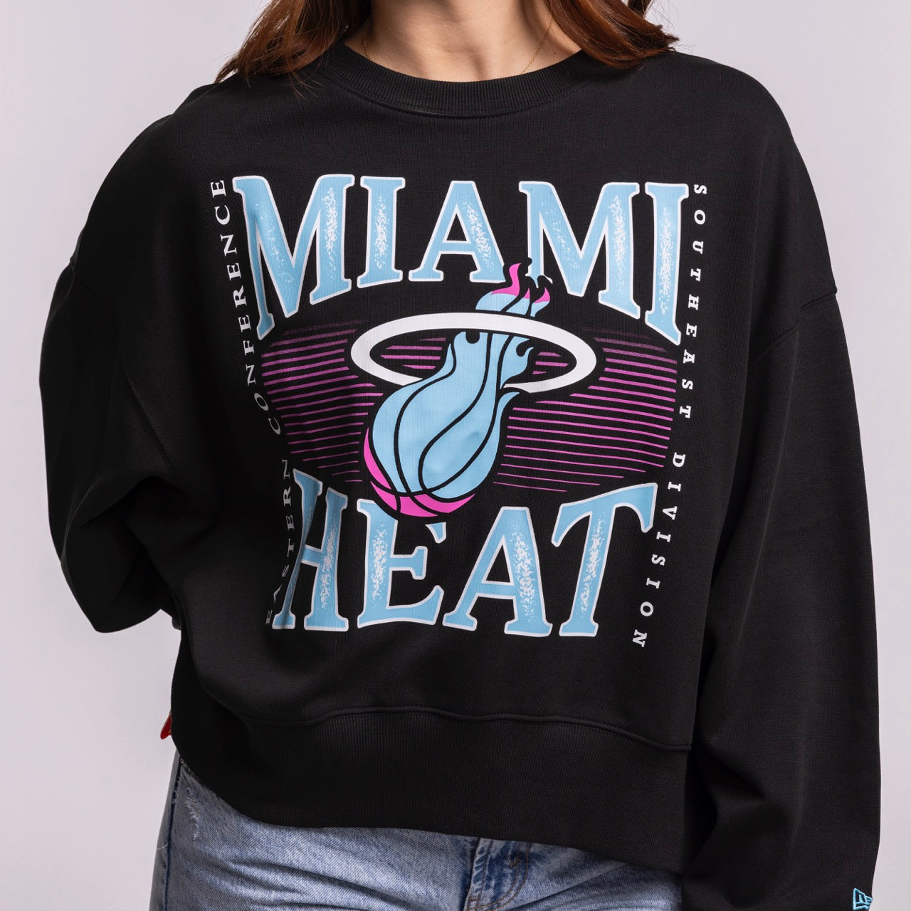 A person wears a size M New Era Miami HEAT Original Vice Nights Wordmark Women's Crewneck in black, featuring a large blue, pink, and white logo. The cotton-polyester sweatshirt obscures their face and is paired with light blue jeans.