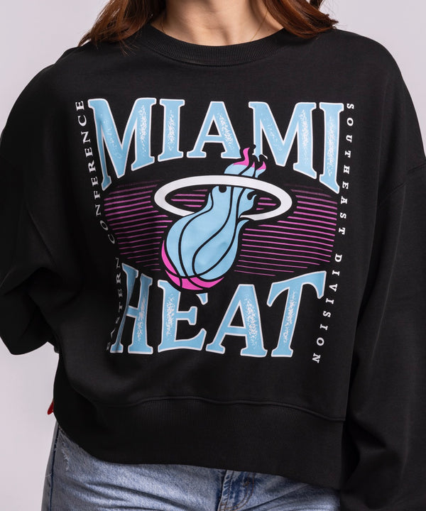 A woman with long brown hair wears the New Era Miami HEAT Original Vice Nights Wordmark Women's Crewneck in black with an oversized cropped fit and light blue jeans, standing against a plain background facing the camera.