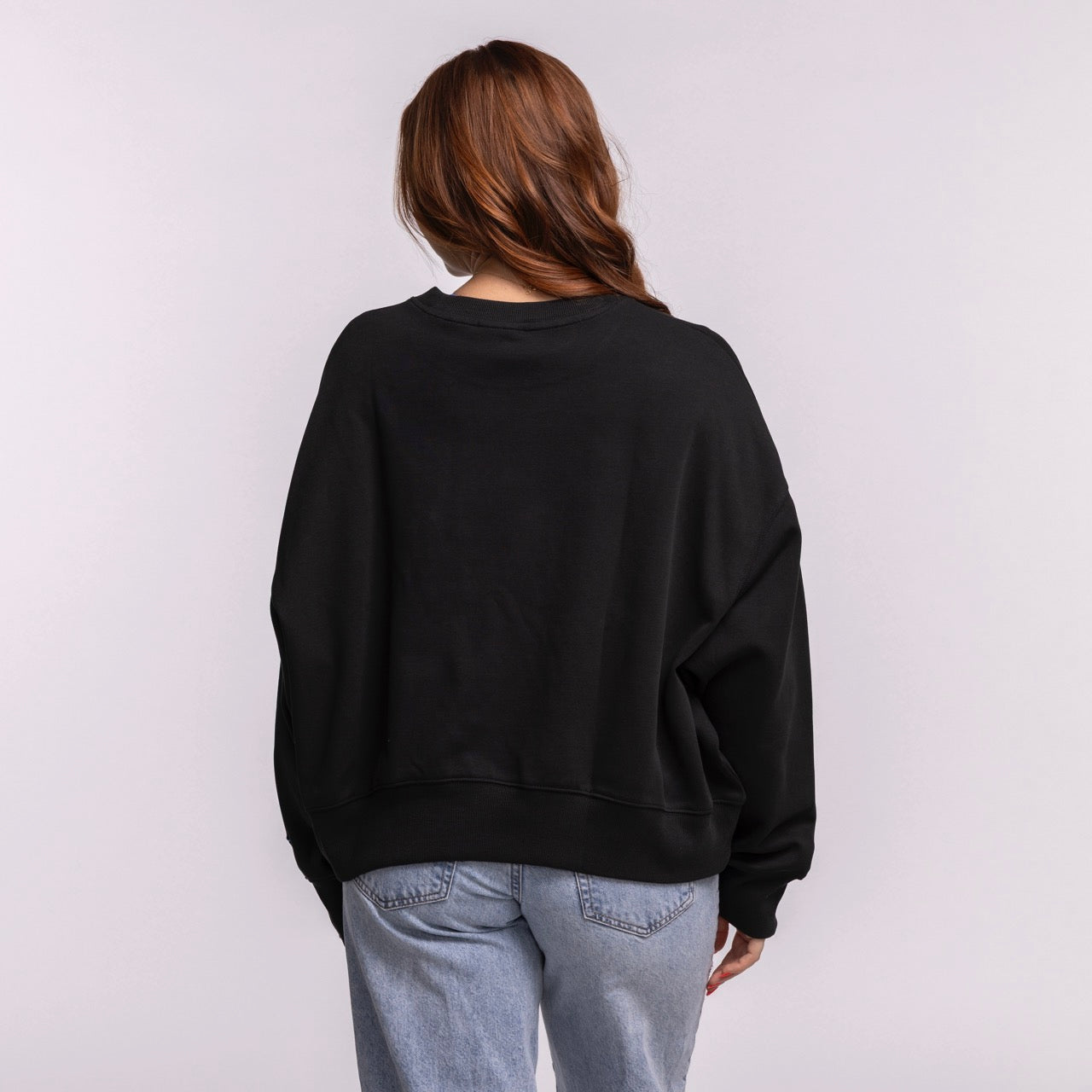 A woman with long brown hair wears the New Era Miami HEAT Original Vice Nights Wordmark Women's Crewneck in size M, styled oversized and cropped with light blue jeans, standing against a plain light background, facing away from the camera.