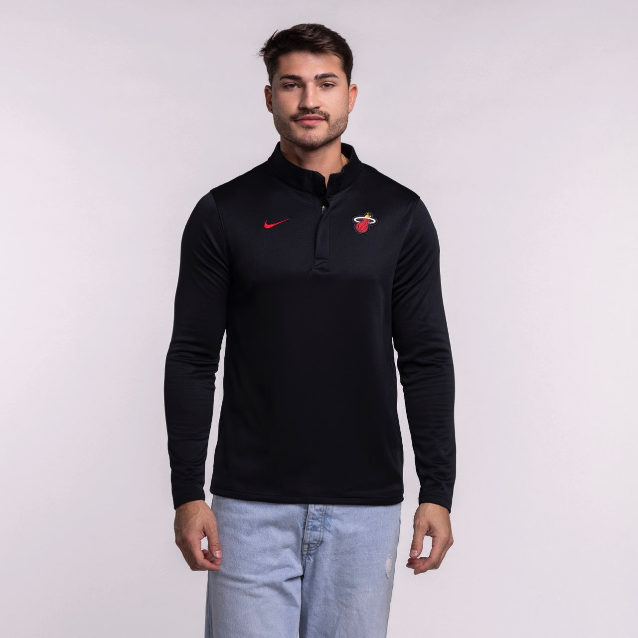 A man with short brown hair and a mustache wears a Nike Miami HEAT Black Logo 1/4 Zip top with a small red Nike logo and team emblem, paired with light blue jeans, standing against a plain light background.